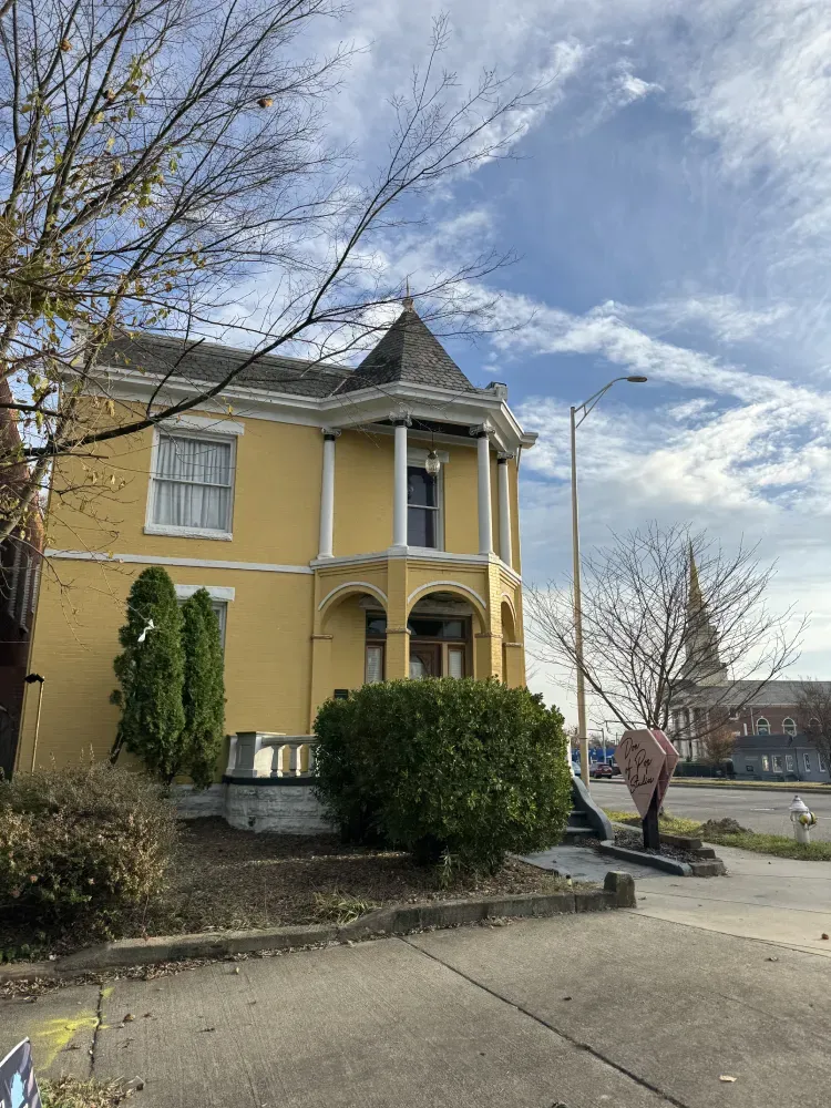 Yellow two-story Victorian house with porch and bushes on a sloped street under a cloudy sky