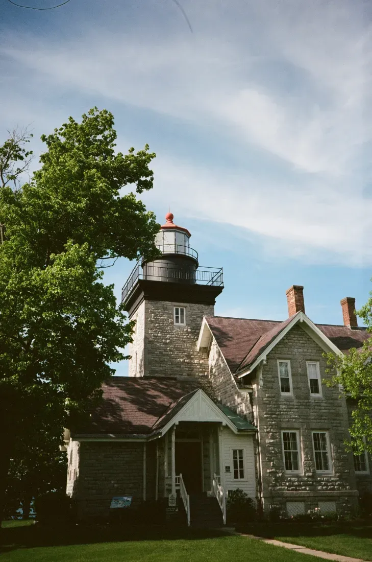 Thirty Mile Point lighthouse beside a gray house under a blue sky, partly framed by green trees