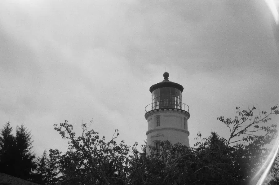 Black and white film photo of the Umpqua River lighthouse against a cloudy sky, trees in the foreground.