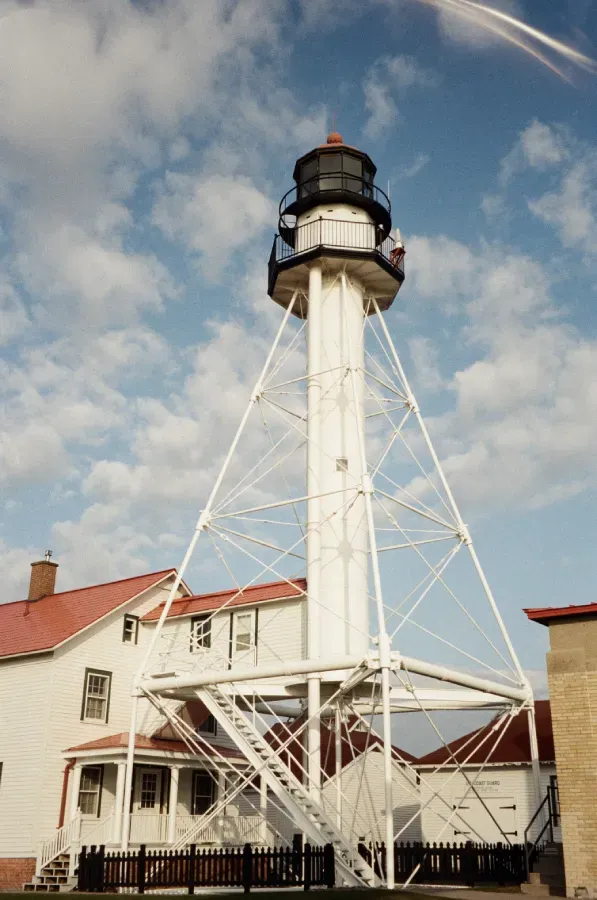 White Fish Point lighthouse with black lantern room, attached to white building with red roof, against a cloudy blue sky.