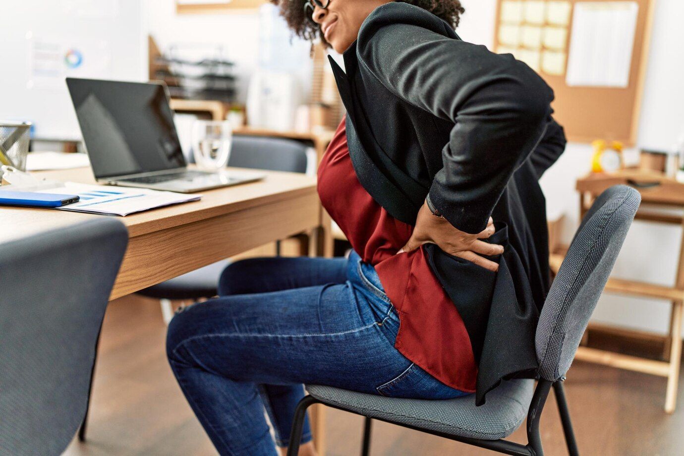 A woman is sitting on a chair holding her back in pain.