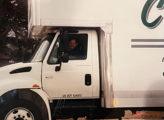 White moving truck with driver visible in the cab, parked outdoors near trees.