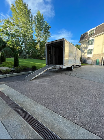 Open trailer with ramp on a paved area beside a green park and building
