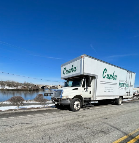 White Cunha moving truck on a snowy road under a clear blue sky, with a stone bridge in the background.