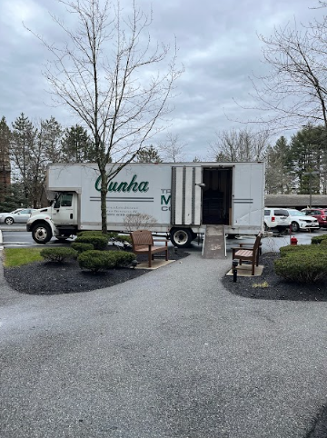 White moving truck with open cargo door parked on a gravel lot near benches and shrubs.