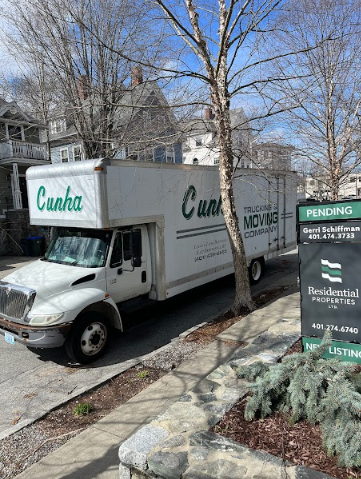 Moving truck parked on a street beside a house and a green real estate sign in a residential neighborhood