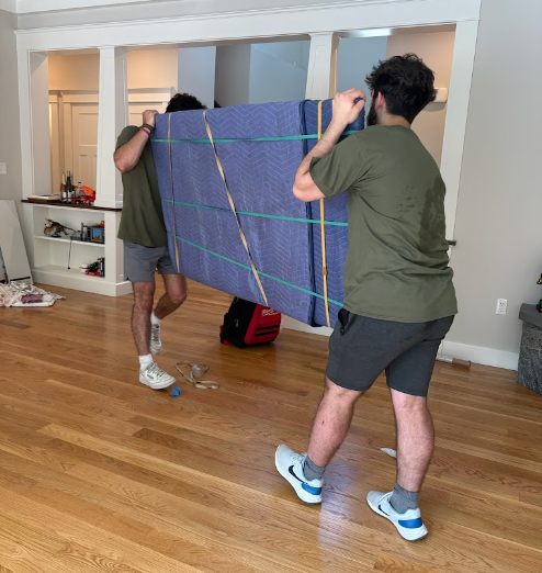 Two people carrying a large blue mattress through a living room