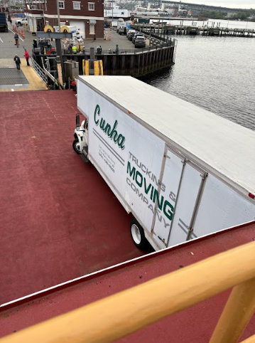 White moving truck on a red ferry deck near a waterfront dock and railing
