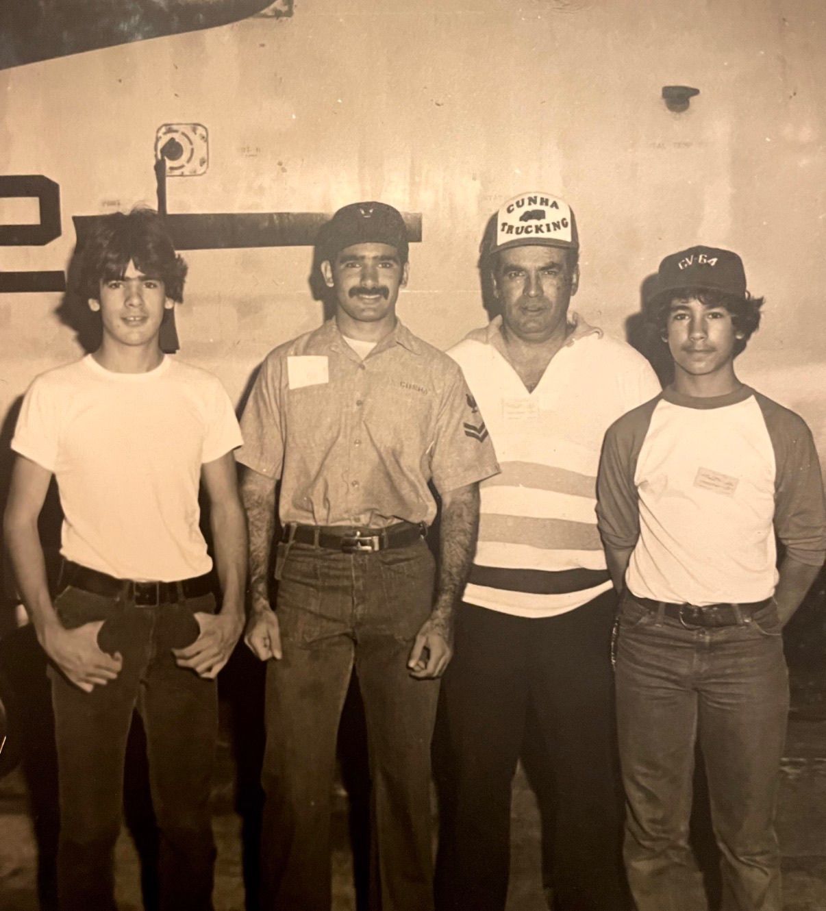 Four workers standing side by side indoors, wearing hard hats and work shirts, posing for a photo.
