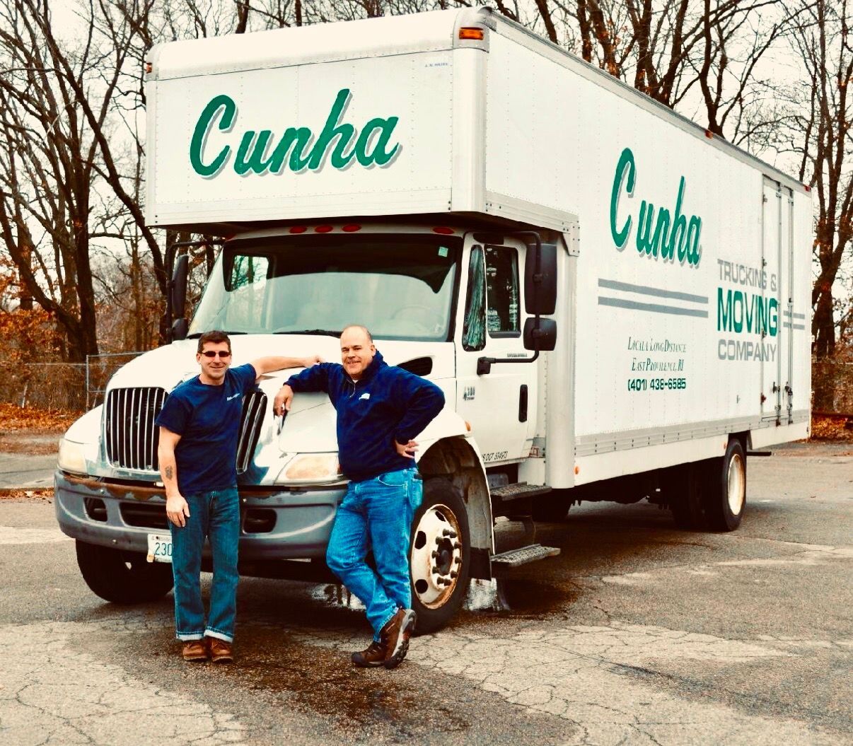 Two men posing beside a white Cunha box truck on a roadside in a wooded area