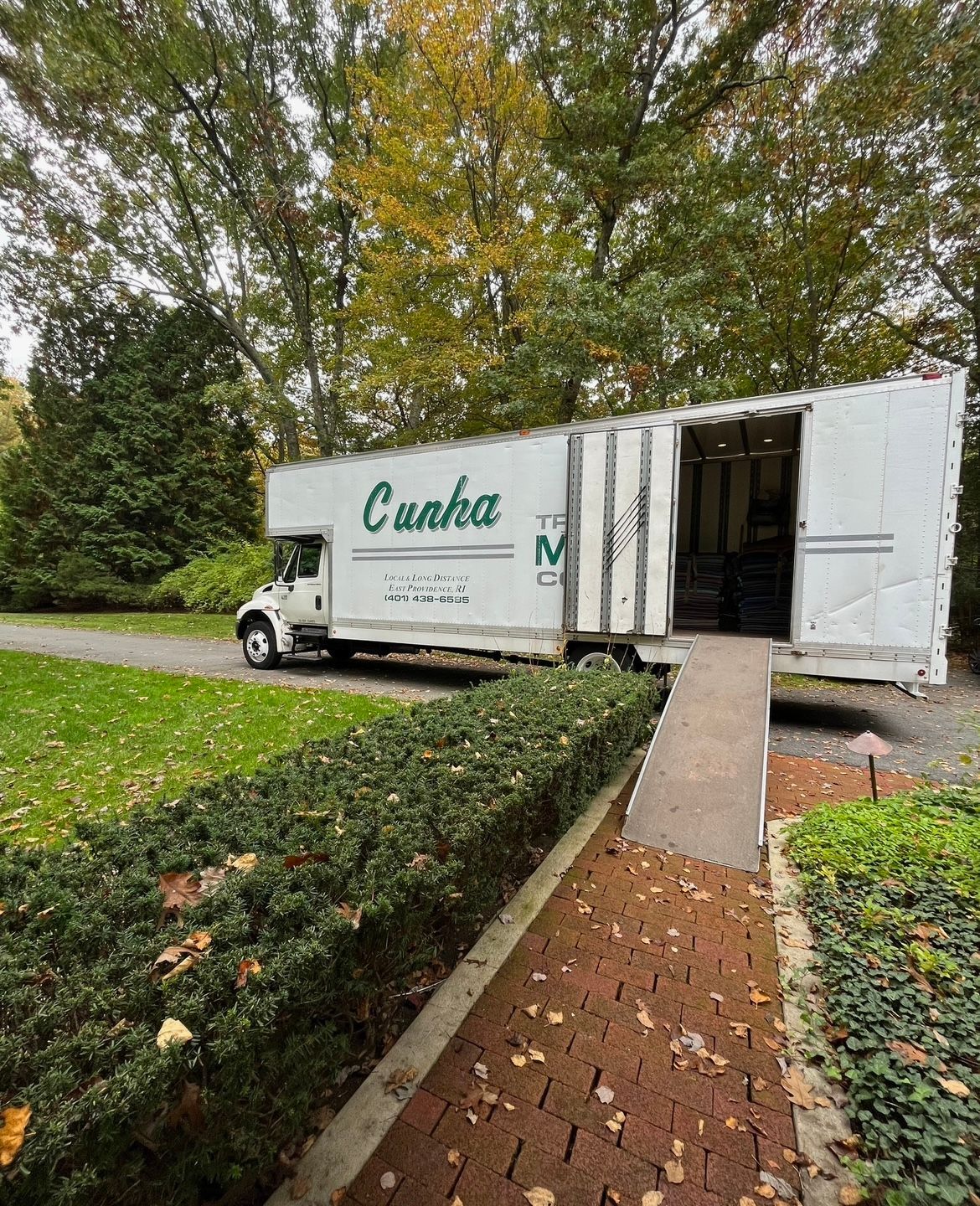 White moving truck with open rear parked beside a brick walkway and hedges on a grassy residential street