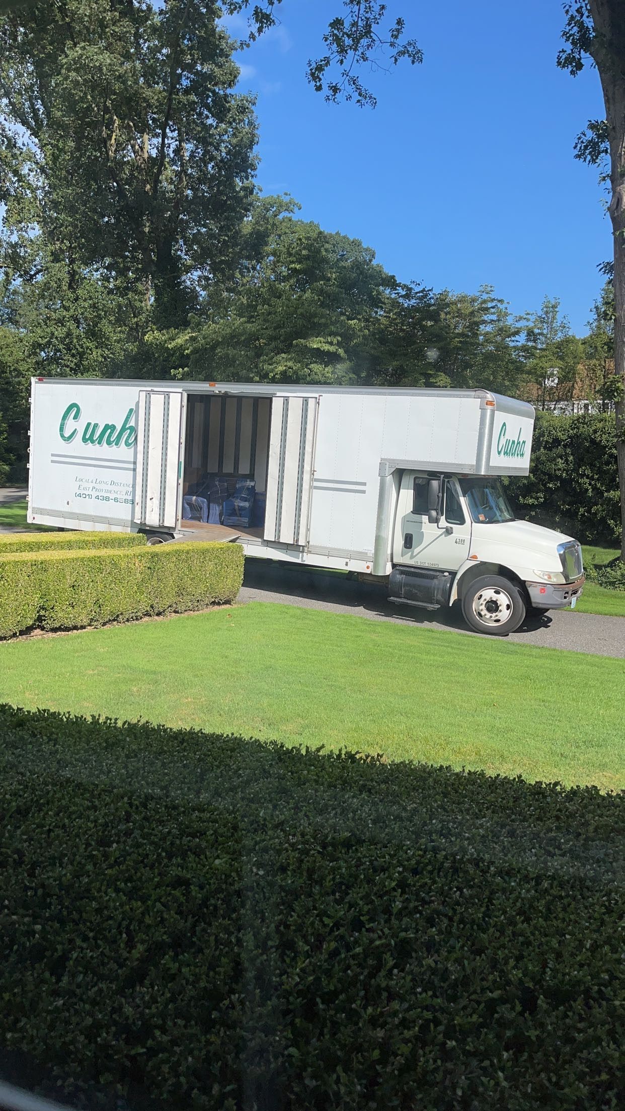 White box truck parked on a grassy driveway beside a hedge and trees under a clear blue sky