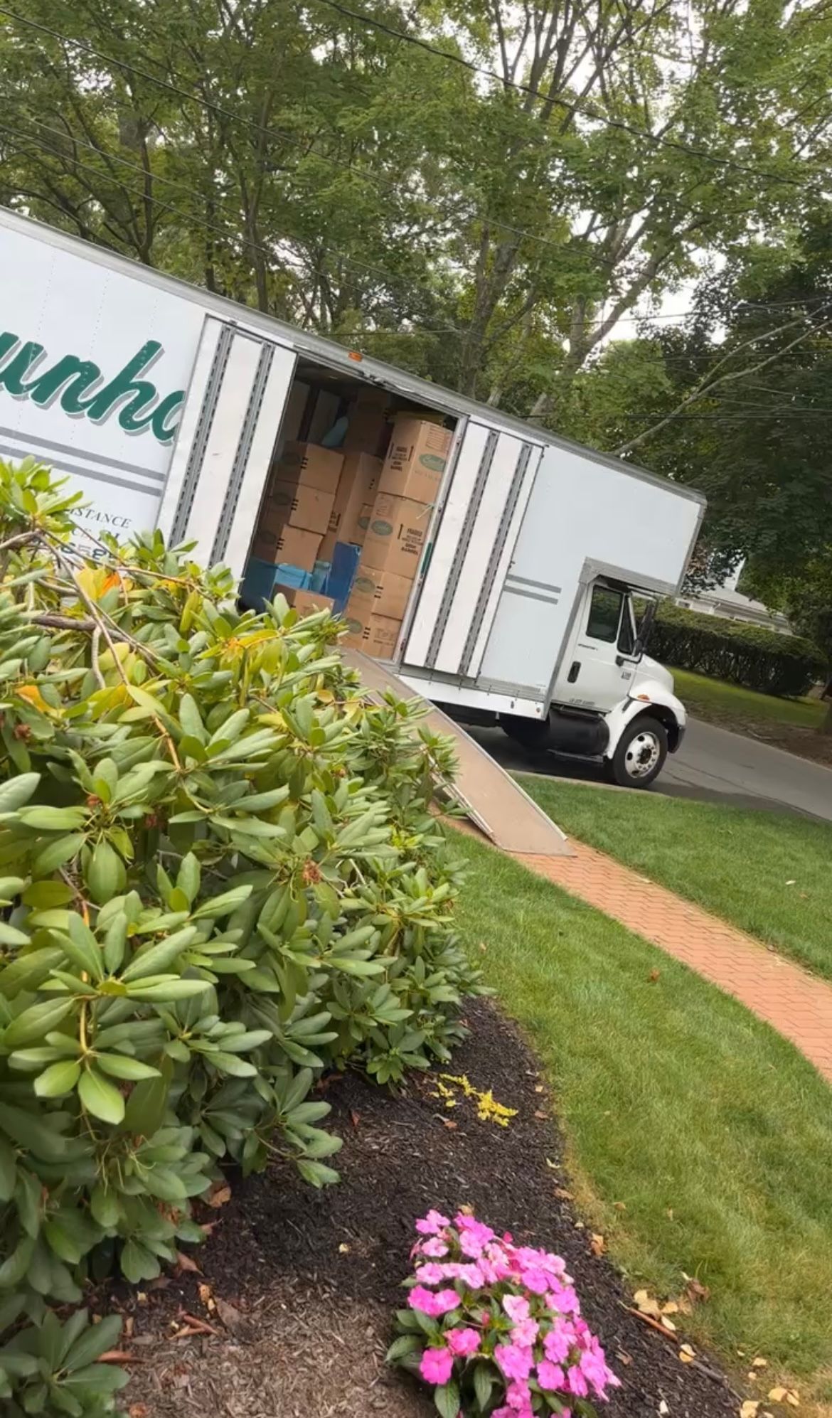 White moving truck with open cargo door beside a landscaped driveway and pink flowers.