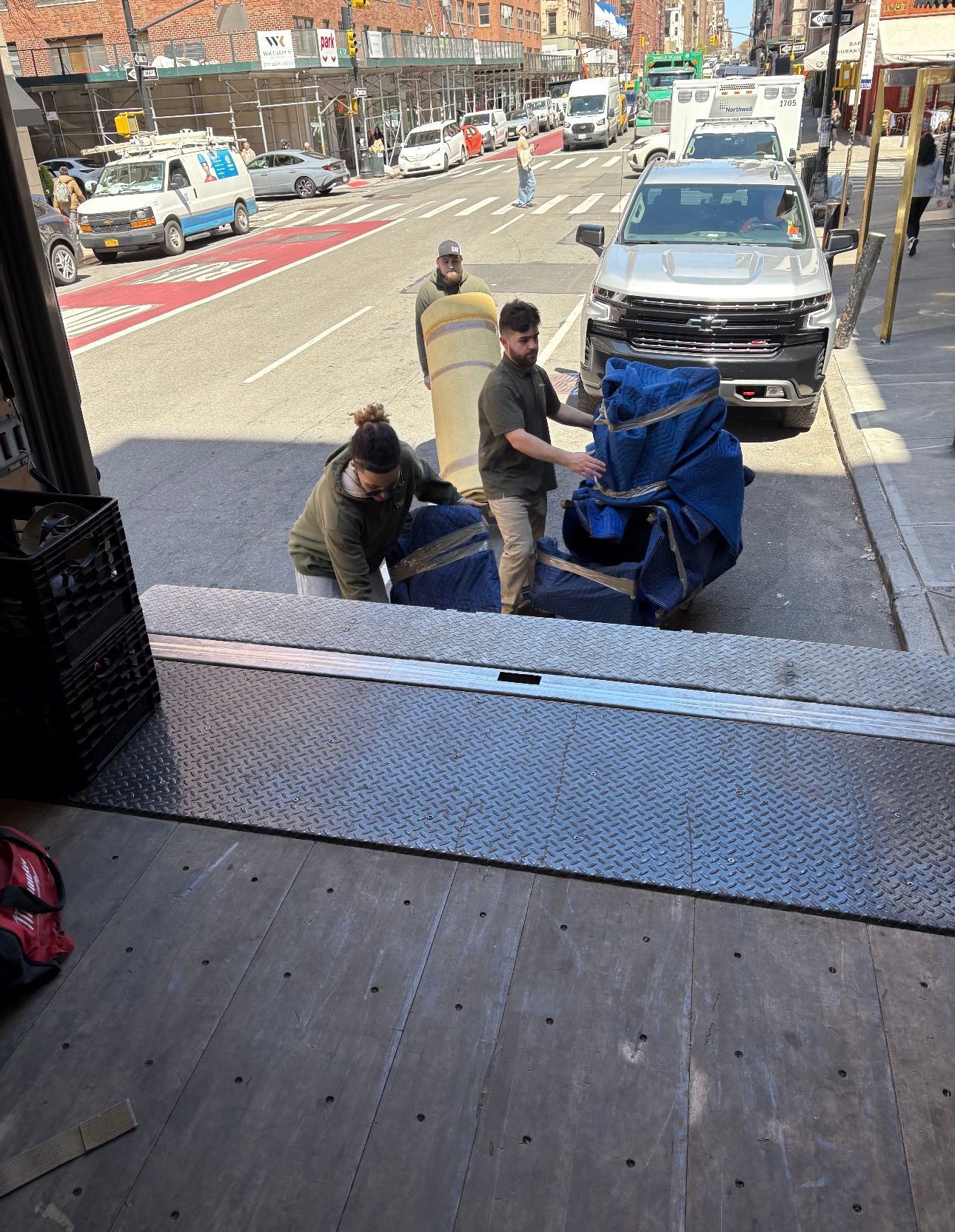 Three people on a truck lift a large blue bag near a curb on a city street.