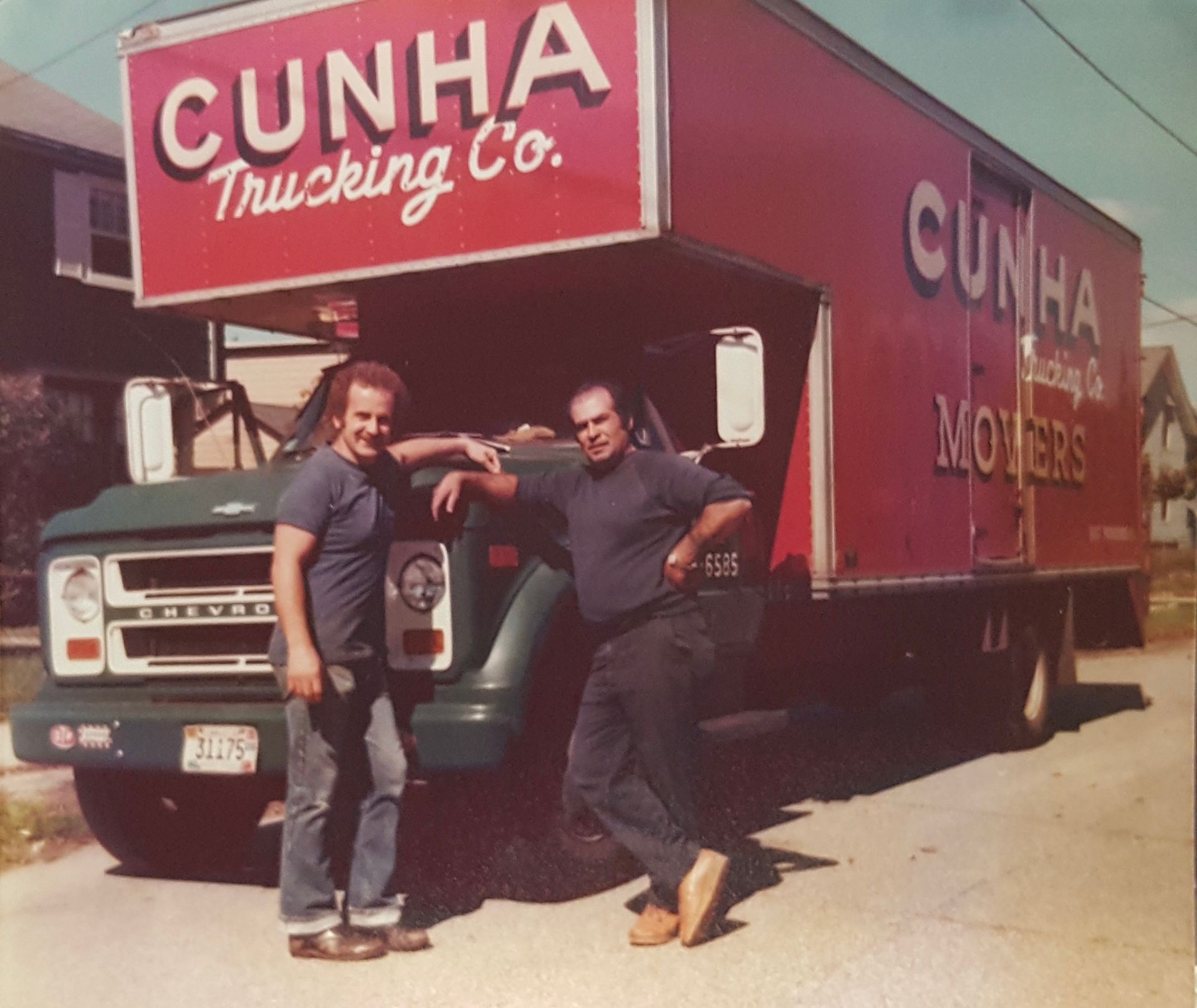 Two men pose beside a red Cunha Trucking Co. semi truck in a sunny lot.