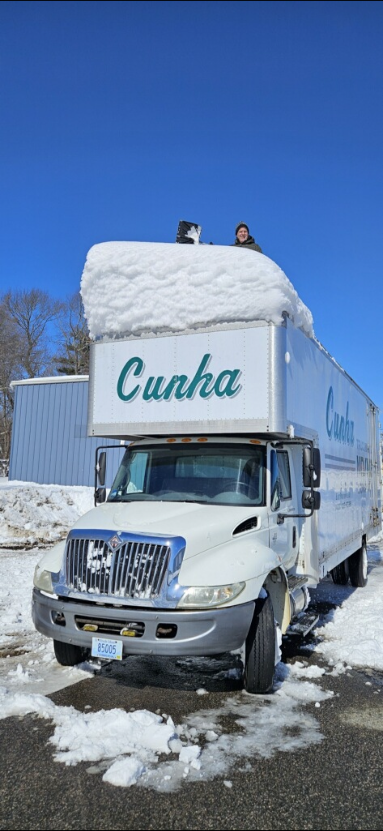 White Cunha box truck buried in snow on a sunny roadside, with two people on top clearing it.