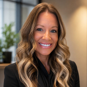 Smiling woman with long wavy hair in a black blazer, standing in a bright office setting