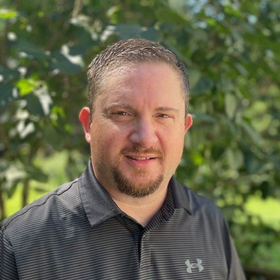 Smiling man in a dark striped polo standing outdoors with green foliage in the background