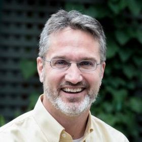 Smiling man with glasses and gray beard outdoors against a leafy background