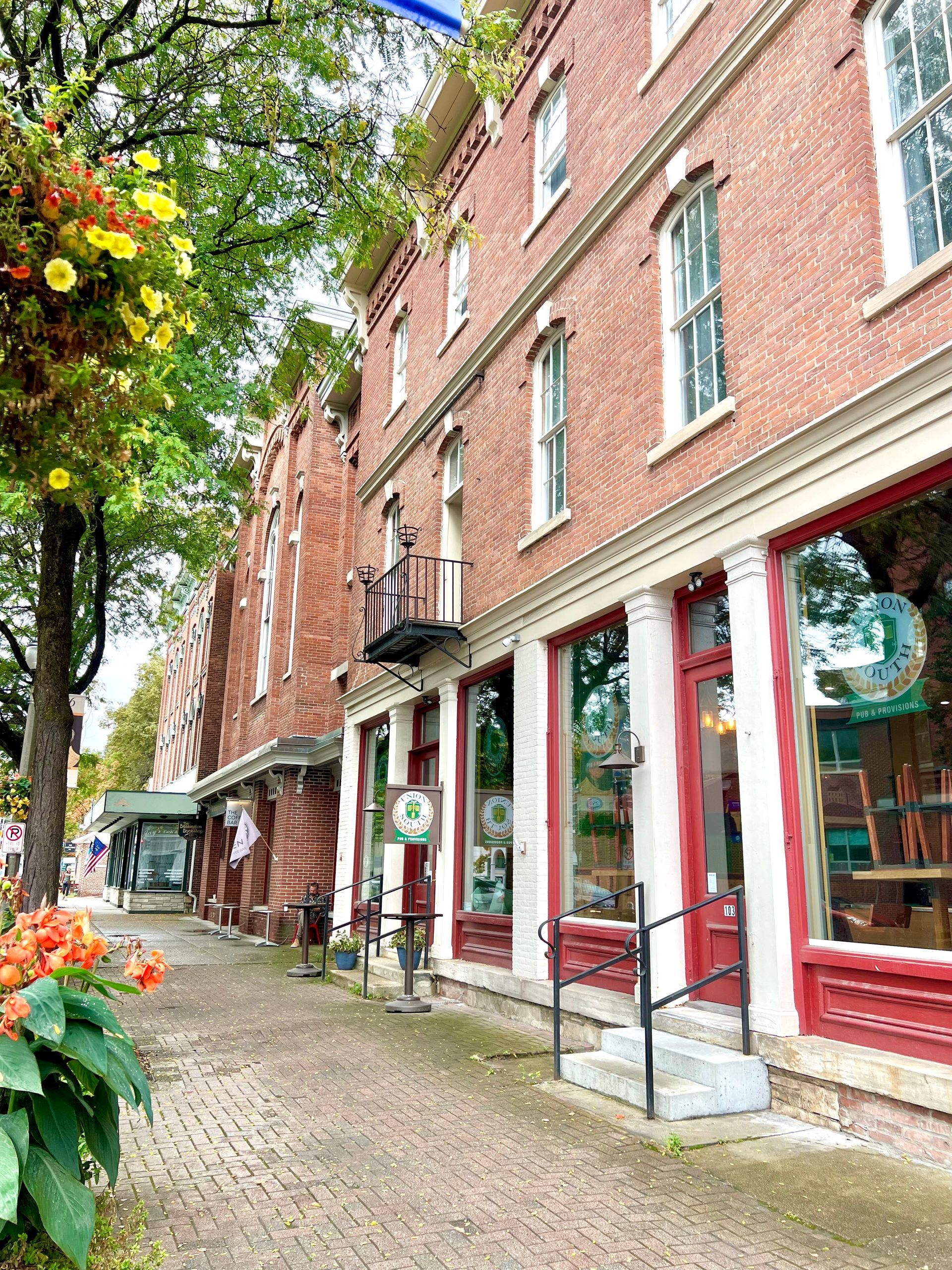 Brick storefronts along a tree-lined cobblestone street with red-trimmed windows and flowers