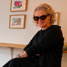 Smiling woman in black clothes and sunglasses seated indoors beside framed wall art
