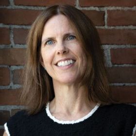 Smiling woman with brown hair in a black sweater standing against a brick wall