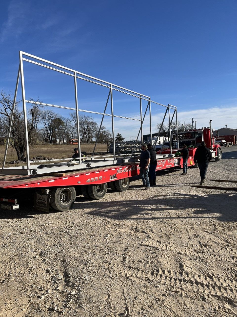 A red semi truck is carrying a large metal structure on a dirt road.