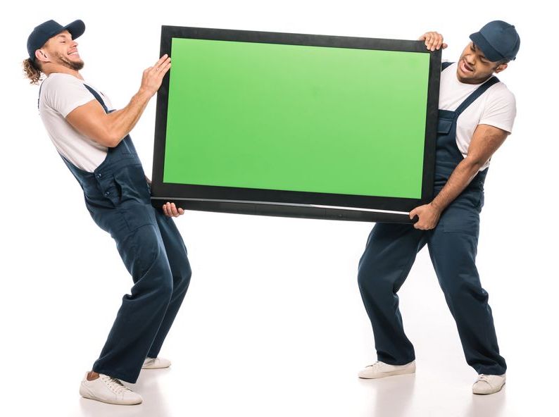 Two men are carrying a flat screen tv with a green screen.