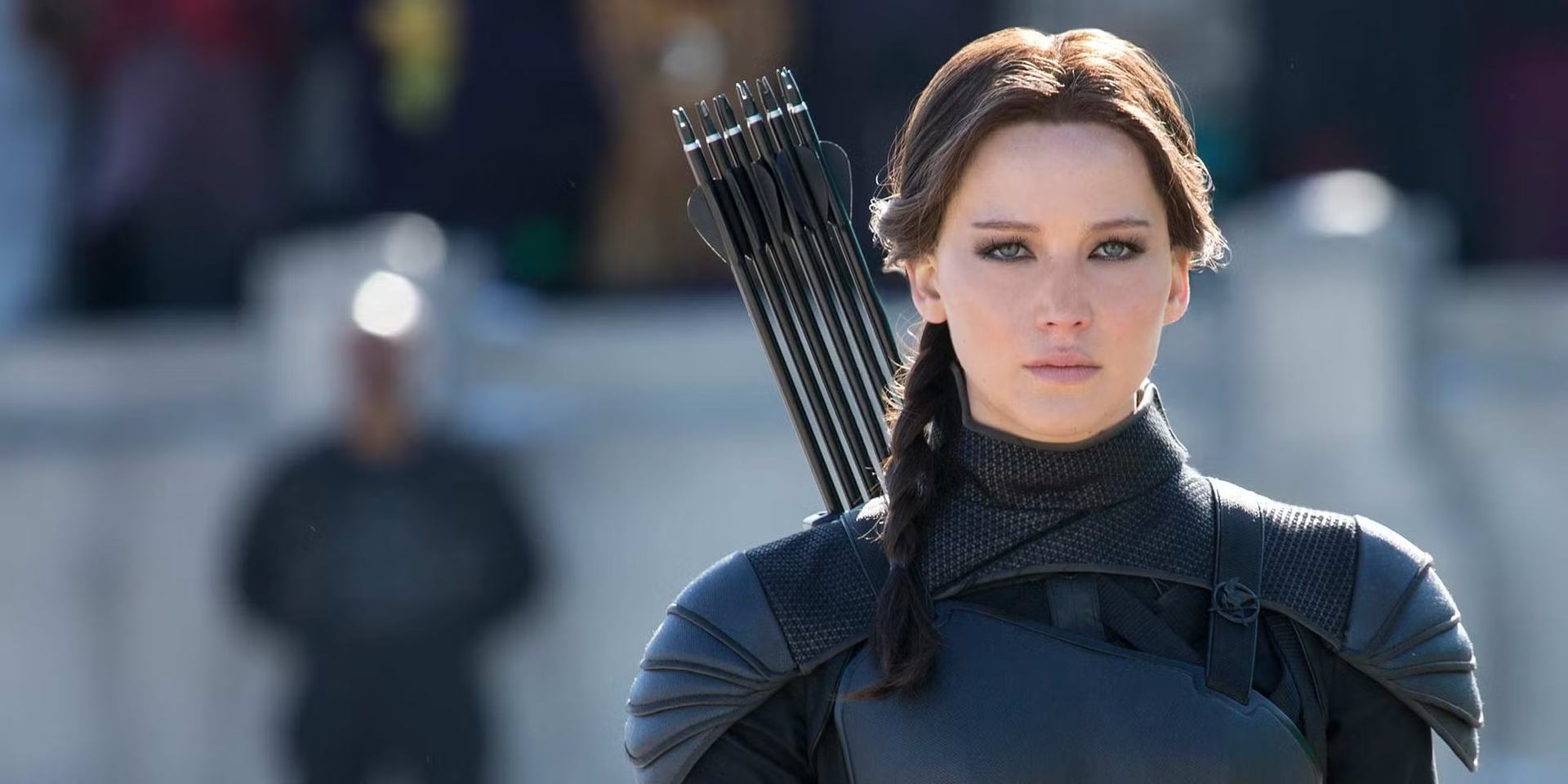 Katniss Everdeen wearing black tactical armor and carrying a quiver of arrows, looking forward with a serious expression.