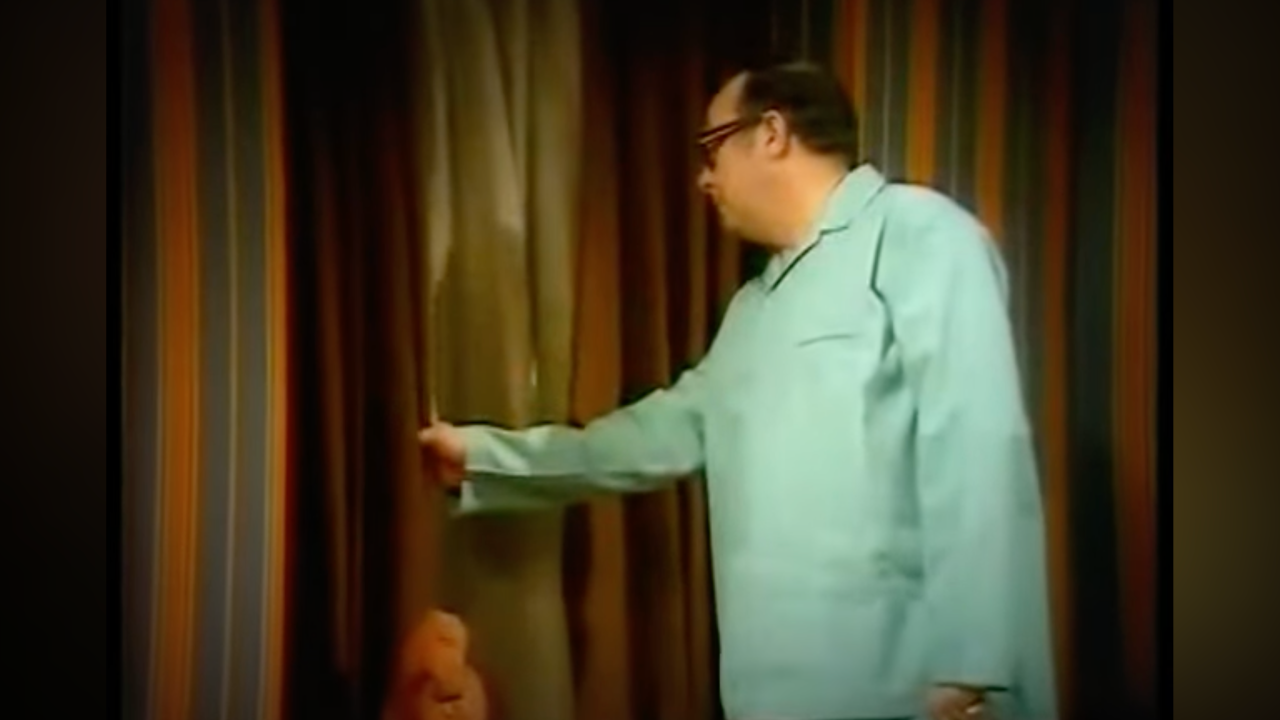 Man in a light blue shirt gesturing toward striped curtains on a stage
