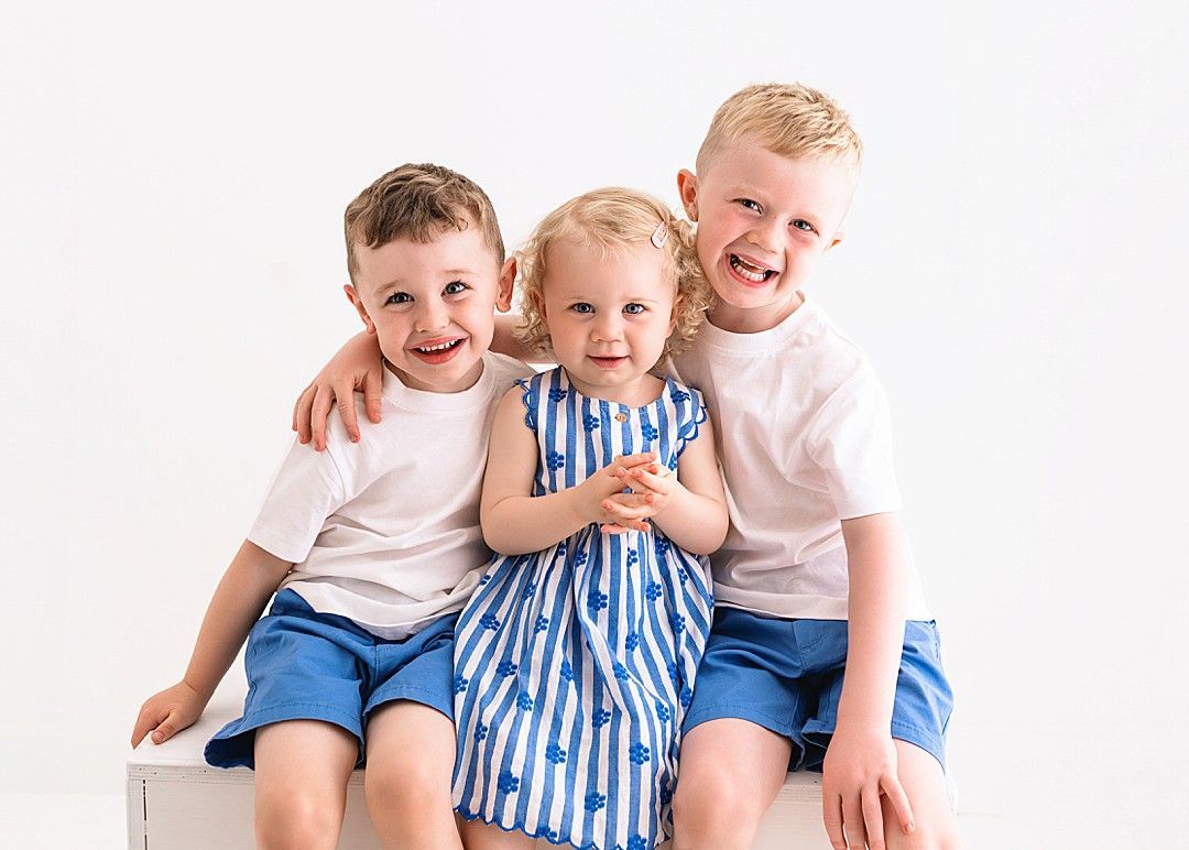 Three smiling siblings sitting together during a relaxed studio family photography session by Dublin family photographer Linda Clarke.