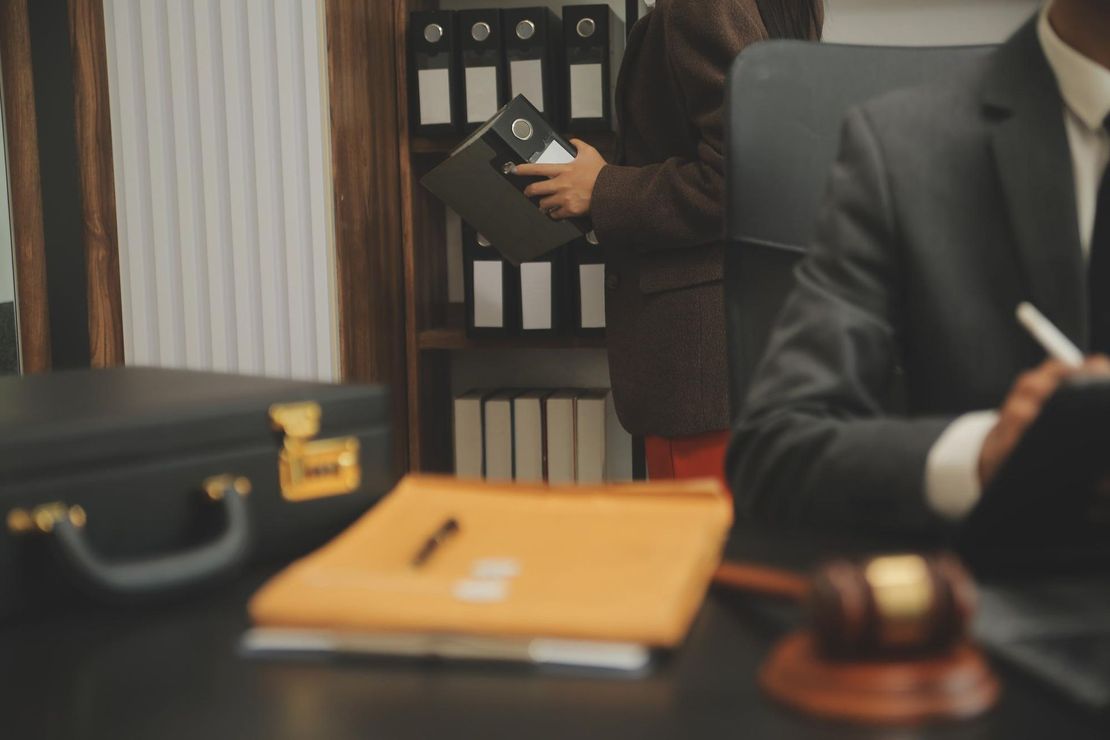 Lawyer’s office desk with briefcase, files, and gavel; two people in suits in the background