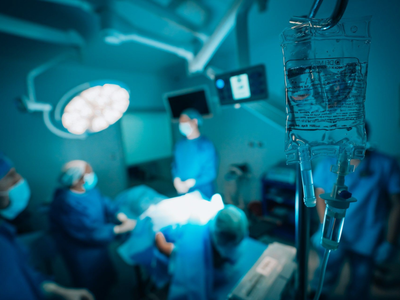 IV bag in foreground with blurred surgeons in an operating room under bright surgical lights