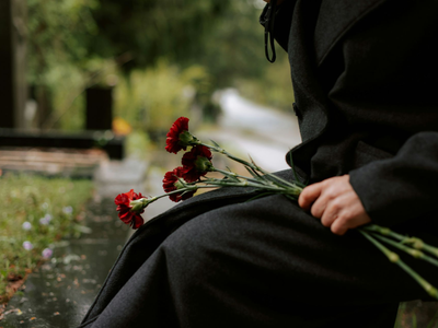 Person in black clothing holding red carnations while seated outdoors near a path.