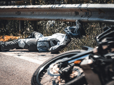 Motorcyclist lying on roadside near guardrail, with a blurred motorcycle in the foreground.