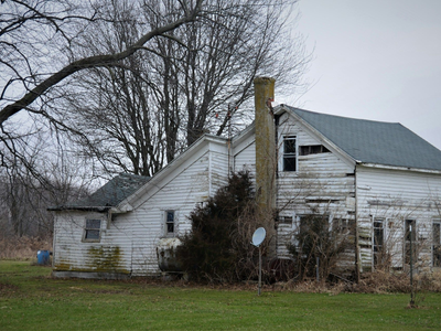 Dilapidated white house with peeling paint, broken windows, and a tall chimney on a gray day