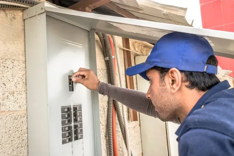 Man in Blue Hat Flipping Circuit Breaker Switch in Electrical Panel — KACE Electrical Solutions In Toowoomba, QLD