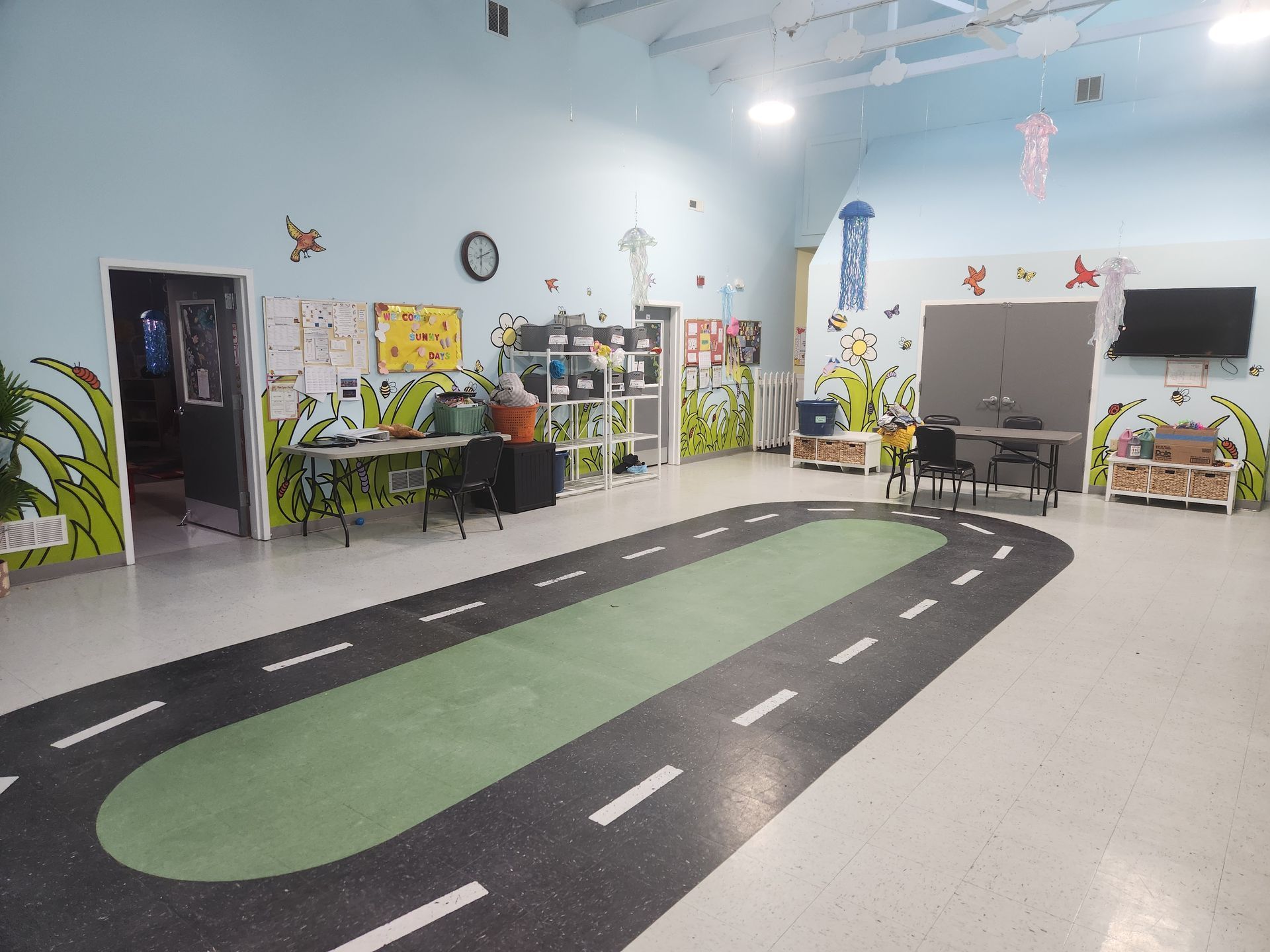 A colorful preschool classroom features a painted oval road track on the floor with tables, chairs, and wall decorations.
