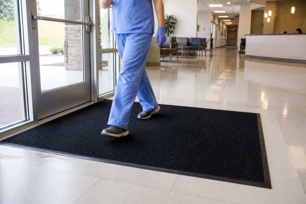 A commercial entrance mat at the doorway of a bright, clean medical office lobby, with a gloved staff member in scrubs walking across it, polished floors visible beyond