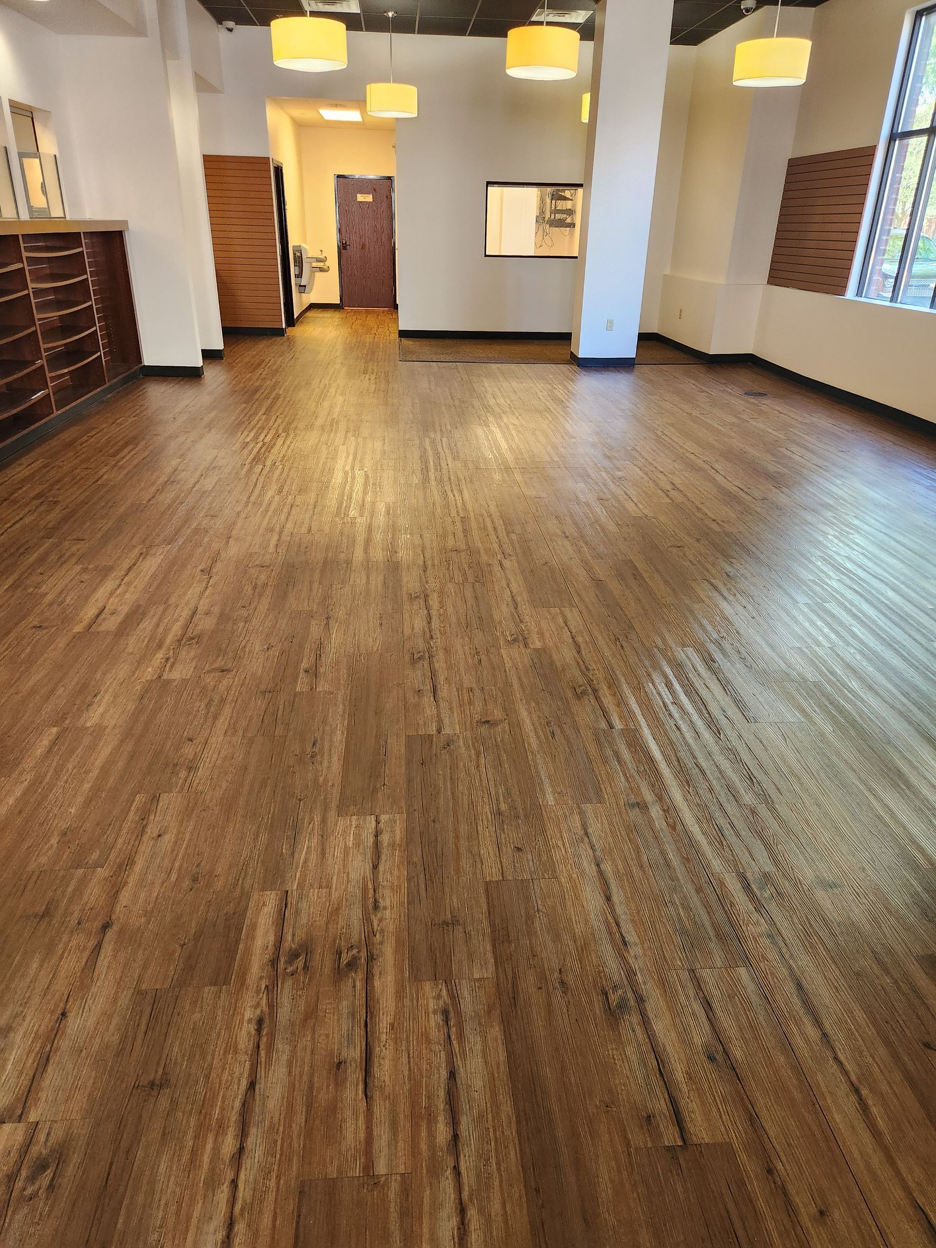 A wide-angle interior view of an empty commercial space with warm-toned wood plank flooring and modern pendant lighting.