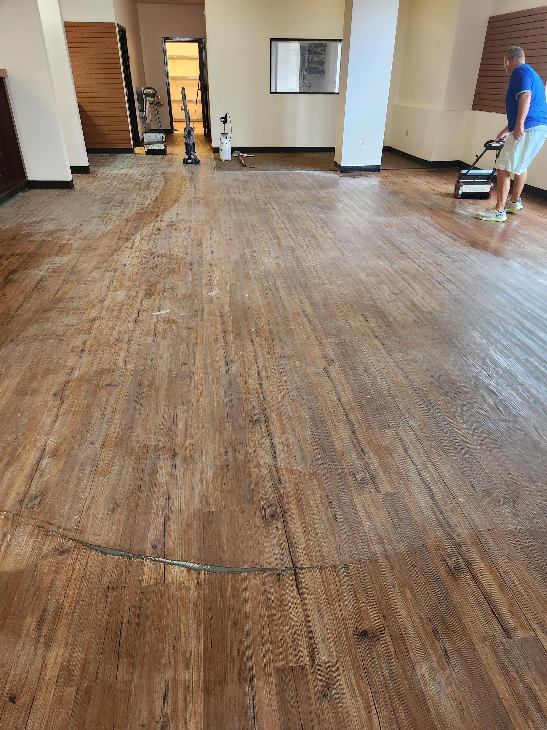 A person in a blue shirt uses a floor buffer on wood-look flooring inside an open commercial space.