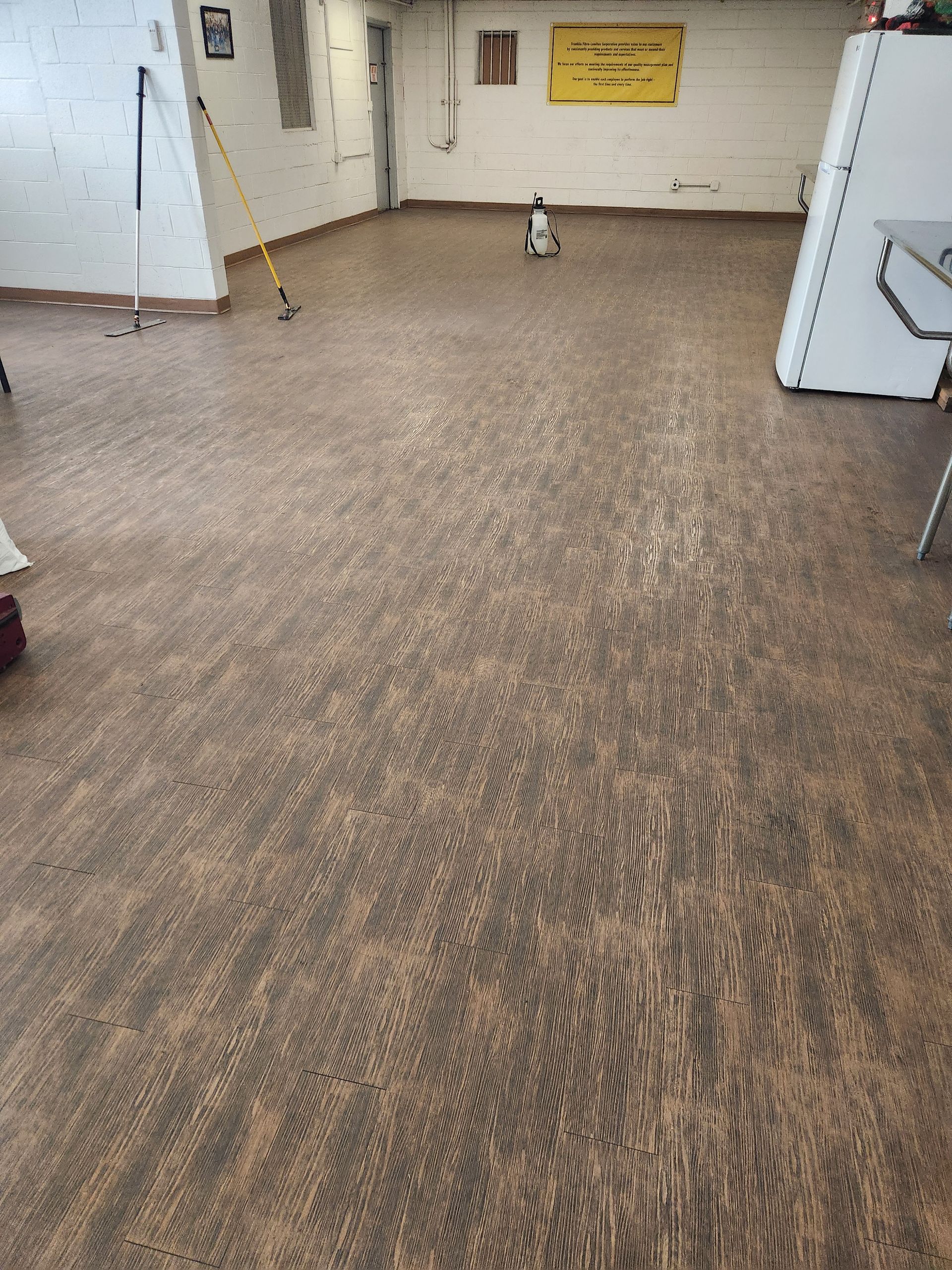 A small robot vacuums a large, open room with wood-patterned flooring, white walls, and a yellow sign.