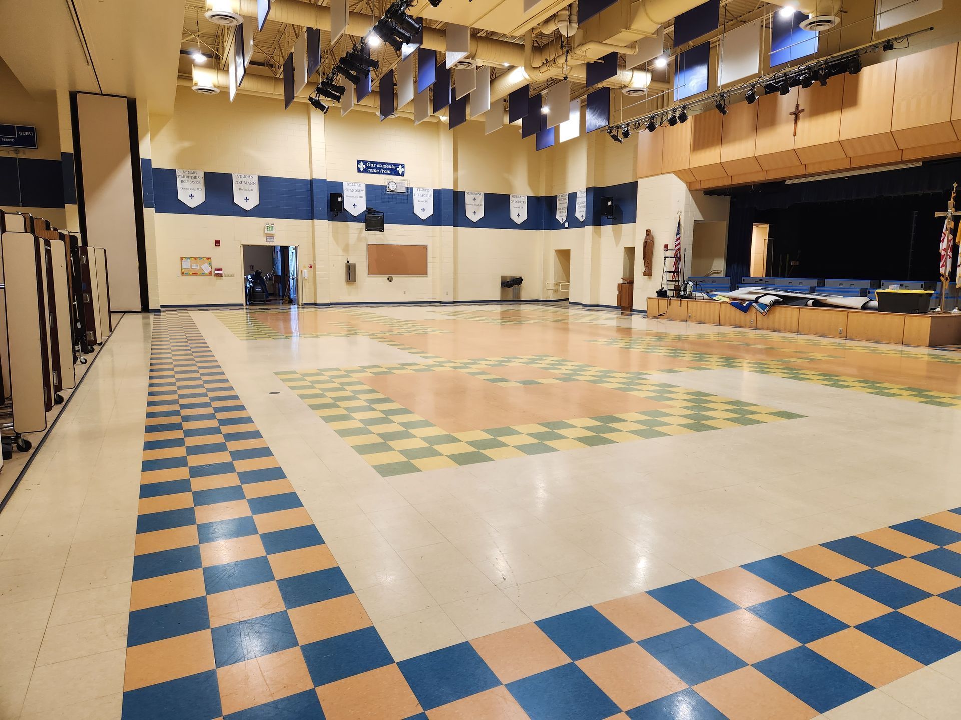 An empty school gymnasium or multi-purpose room with checkerboard-patterned floor borders and a stage at the far end.