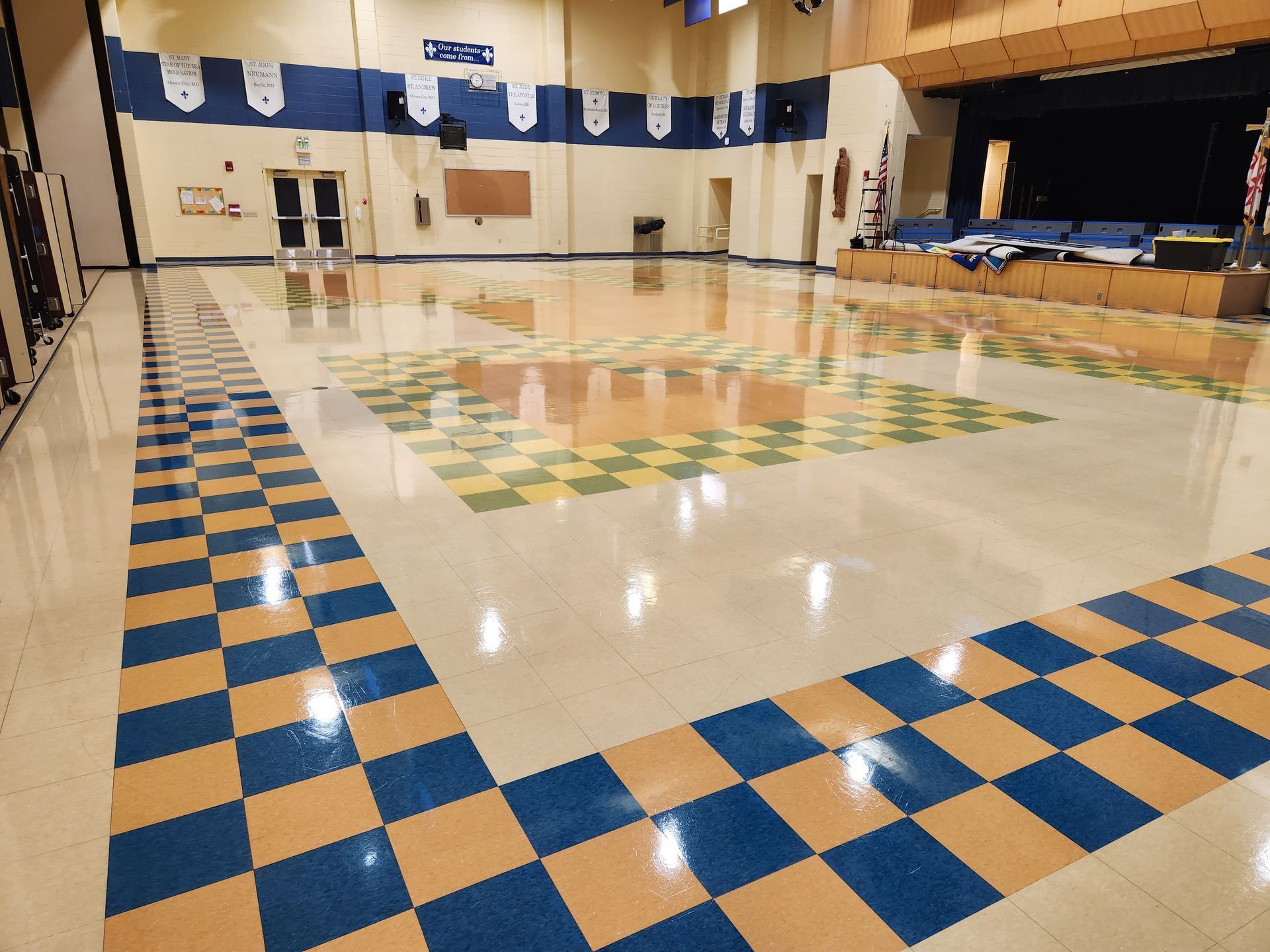 An empty school gymnasium with polished, checkerboard-patterned tile floors, blue and gold accents, and a raised stage.