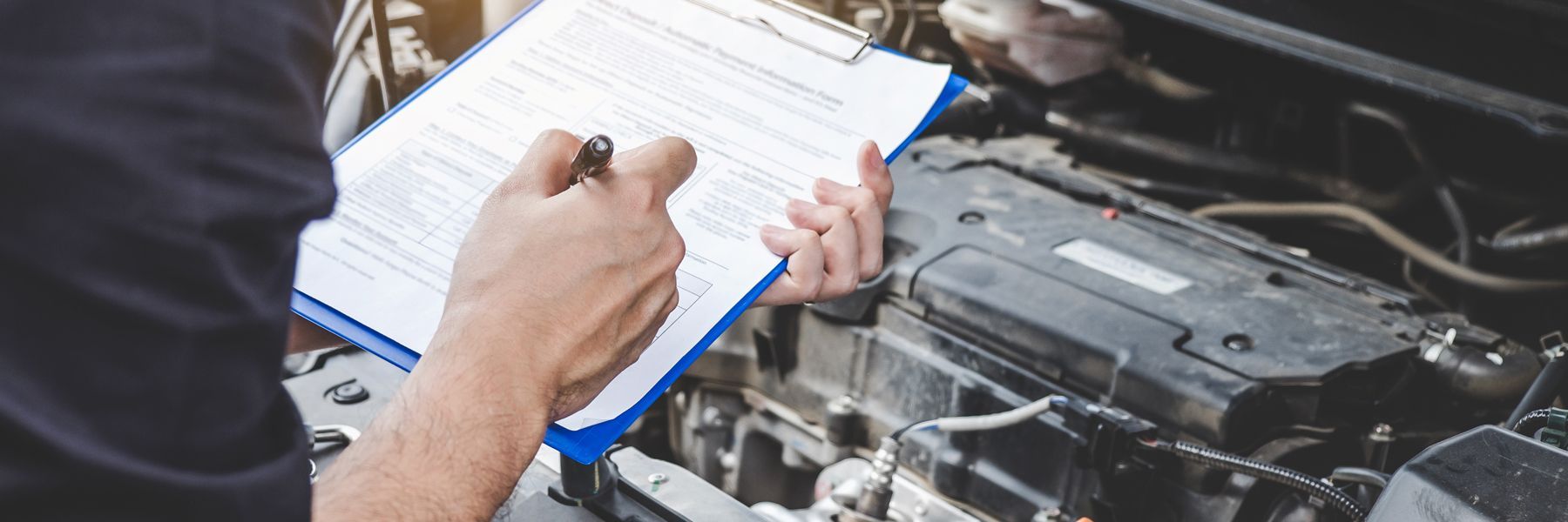 A man is writing on a clipboard while working on a car engine.