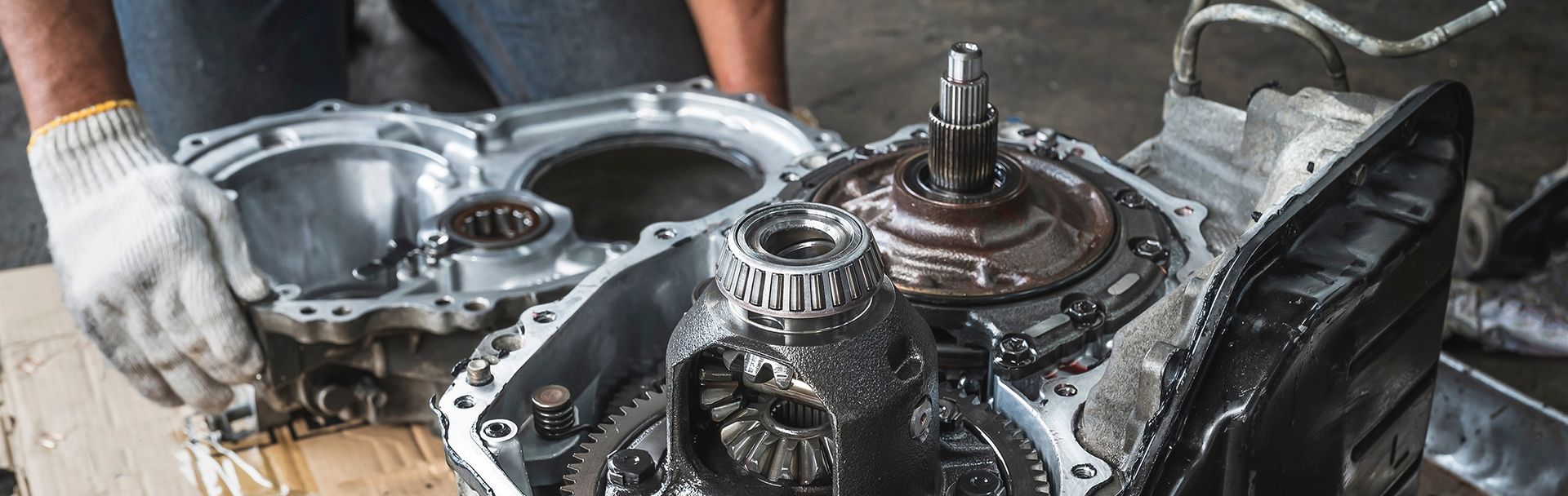 Mechanic working on a car transmission, holding a part, gloves, gears, and oily components are visible.