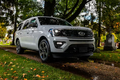 White Ford Expedition SUV parked on a gravel path in a cemetery, surrounded by trees and headstones.