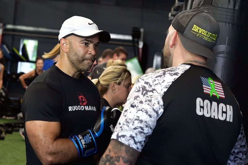 Man in white cap talks with coach in black shirt at Revolution Fitness in East Northport, NY both standing face-to-face.