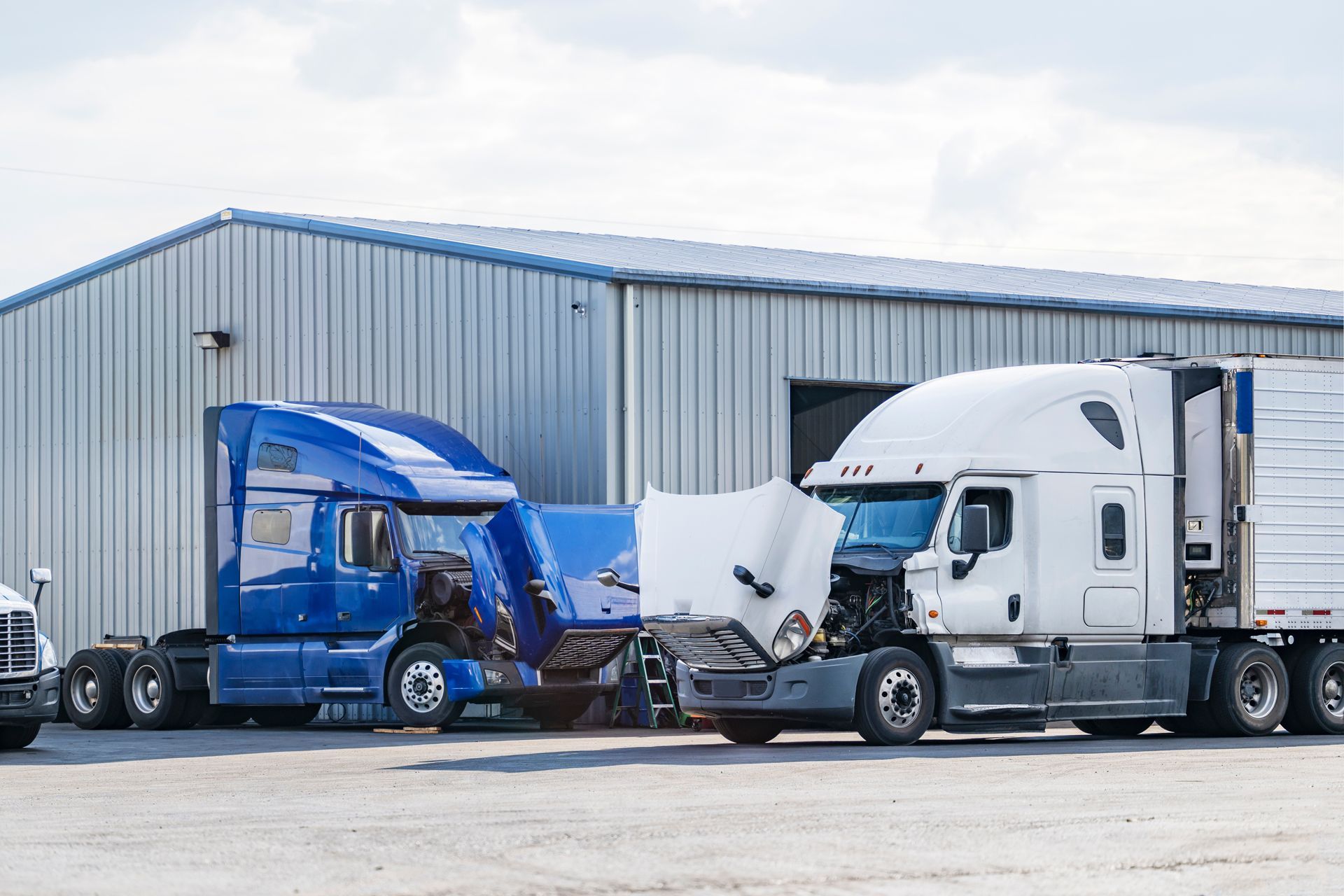 Blue and white semi-trucks with hoods open in front of a metal building