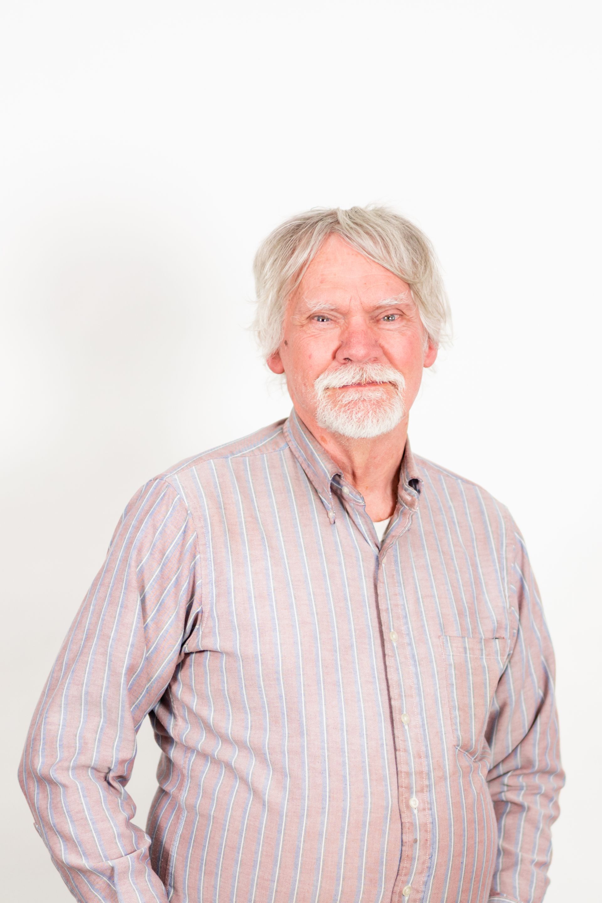 An older man with gray hair and mustache smiles, wearing a striped shirt, against a white backdrop.